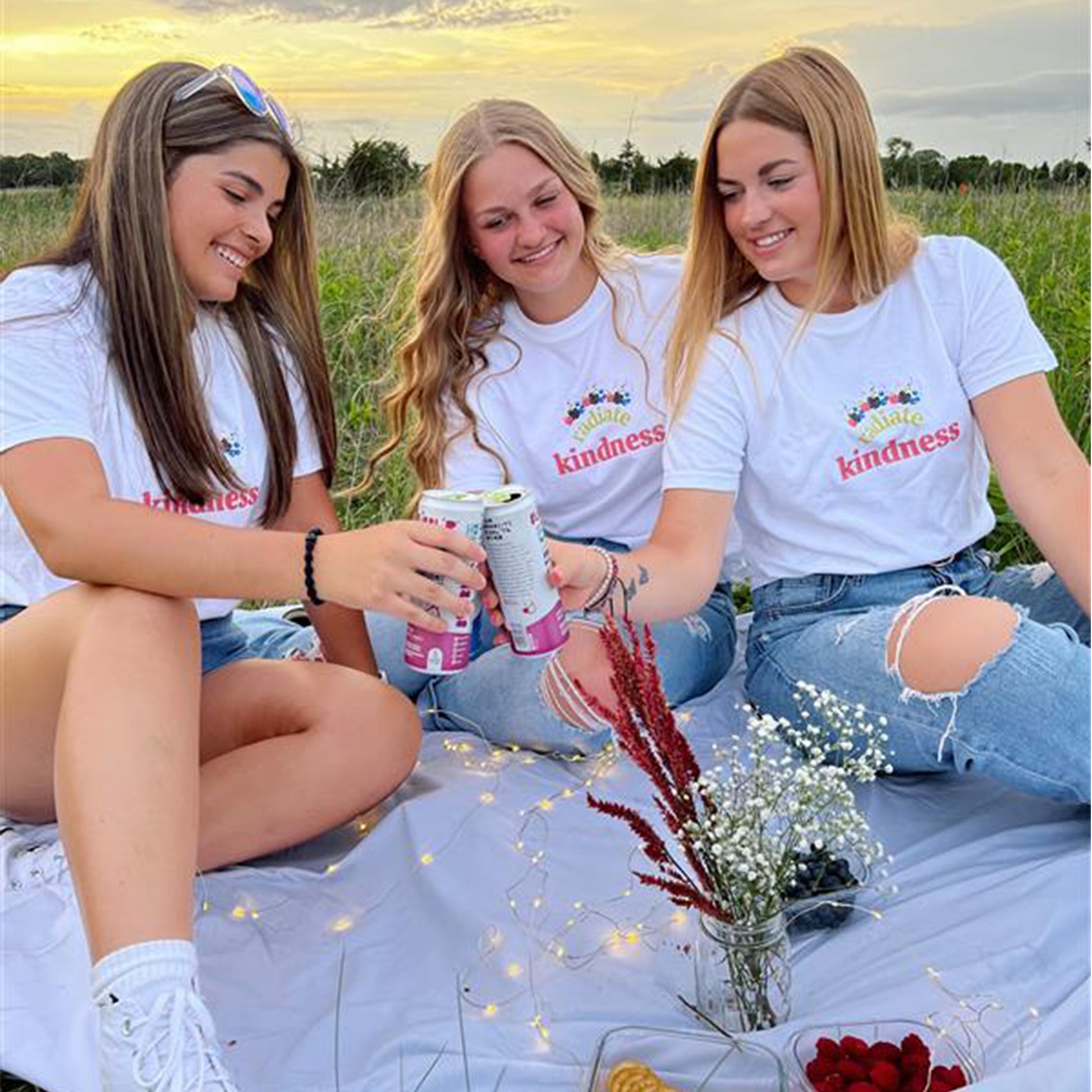 three young women toasting with three bubbl'r cans wearing white radiate kindness tees